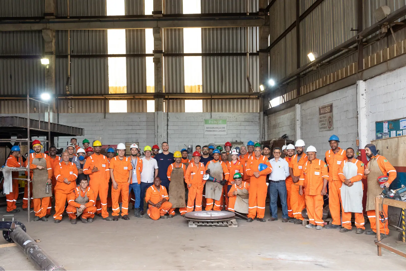 Equipe AASJ — trabalhadores em EPIs laranja reunidos no galpão industrial